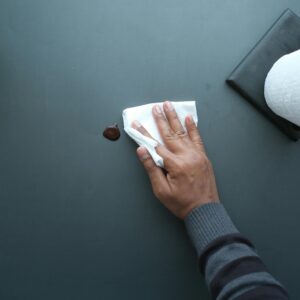 A hand wipes a dark surface with a tissue near a paper roll and plant.