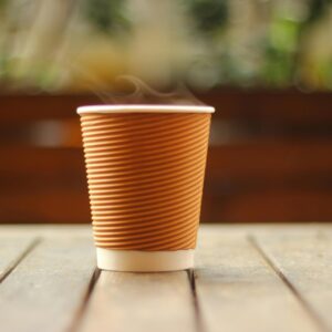 A close-up of a steaming hot coffee in a brown paper cup on a wooden table indoors.