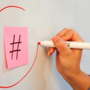 Close-up of hand drawing a circle around a hashtag on a sticky note with a red marker.