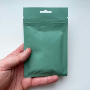 A green resealable pouch held by a hand on a neutral backdrop.