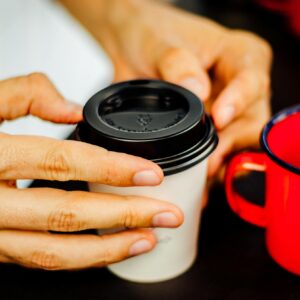 A close-up image of hands holding a disposable coffee cup with a plastic lid next to a red mug.