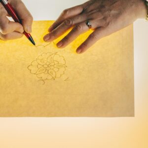 Hands tracing a floral design on backlit paper, showcasing artistic craftsmanship.