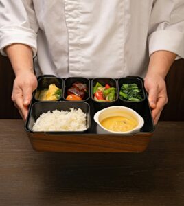 Chef holding a bento box with rice, vegetables, and soup, showcasing traditional Japanese cuisine.