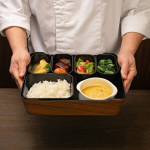 Chef holding a bento box with rice, vegetables, and soup, showcasing traditional Japanese cuisine.