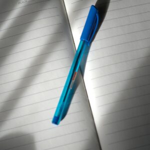 A lined notebook open with a blue pen placed on it, lit by natural light.