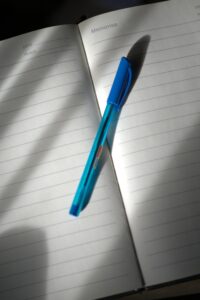 A lined notebook open with a blue pen placed on it, lit by natural light.