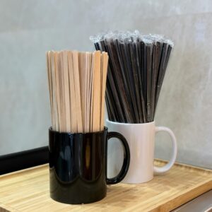 Wooden coffee stirrers and wrapped black straws in mugs on a bamboo tray, indoor cafe setting.