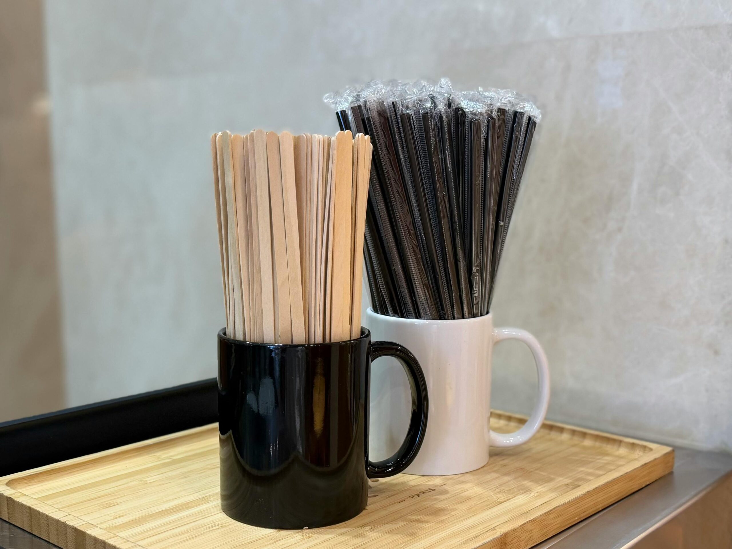 pexels photo 36894279 36894279 1 Wooden coffee stirrers and wrapped black straws in mugs on a bamboo tray, indoor cafe setting.
