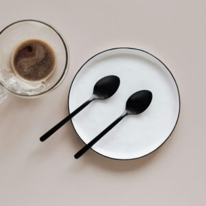 From above composition of ceramic plate with black spoons placed near glass cup of coffee on beige table