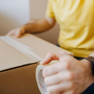 Crop anonymous male in casual wear using cling tape to seal cardboard container with belongings during relocation