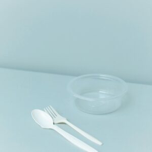 An empty clear plastic container with a spoon and fork on a soft pastel background.