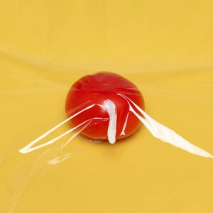 A fresh tomato wrapped in clear plastic wrap on a vibrant yellow background, highlighting freshness and packaging.