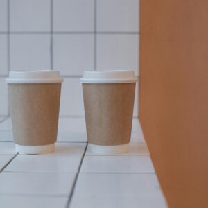 Two disposable coffee cups on a white tiled surface against an orange wall, minimalist style.