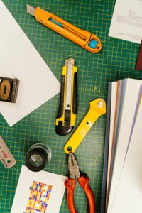 Top view of various cutting tools on a green grid mat surrounded by papers, ideal for creative projects.