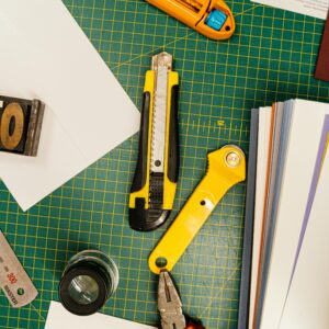 Top view of various cutting tools on a green grid mat surrounded by papers, ideal for creative projects.