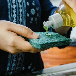 Person pouring green dishwashing soap onto a scrub pad in a kitchen setting.