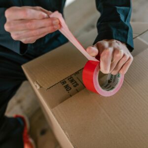 Focus on hands sealing a cardboard box with red tape, depicting careful packaging.