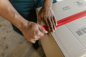 Close-up of hands sealing a cardboard box with red tape indoors.