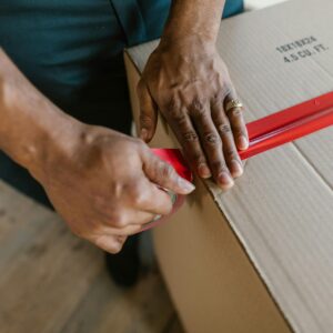 Close-up of hands sealing a cardboard box with red tape indoors.