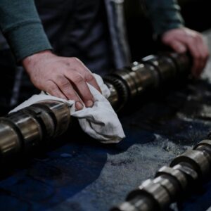 Close-up of a mechanic cleaning a camshaft with a cloth in an industrial setting.