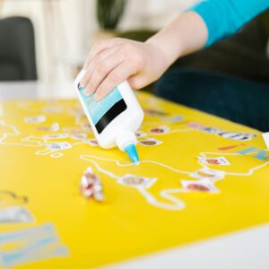 Close-up of hands using glue for an art project on yellow paper with cutouts.