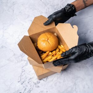 Delicious burger and seasoned fries in a cardboard takeout box, held by gloved hands.