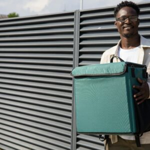 African American man smiling while holding a thermal bag outdoors near a modern fence.