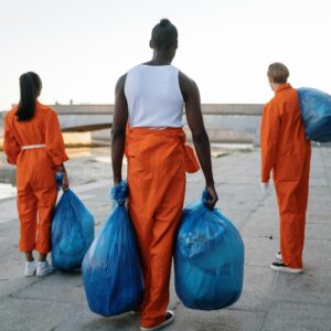 Three people in orange overalls collecting garbage by a river as part of an urban community service project.